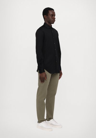 Svart langermet skjorte med knappdesign, kombinert med olivengrønne bukser og hvite joggesko, stående mot en ensfarget bakgrunn.