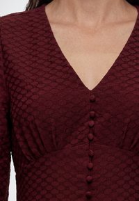 Gros plan d'une robe bordeaux texturée avec un décolleté en V et des boutons décoratifs recouverts de tissu sur le devant.