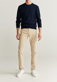 Muž v námořnickém dlouhém rukávu, béžových slim-fit kalhotách a bílých teniskách stojí před jednobarevným světlým pozadím.