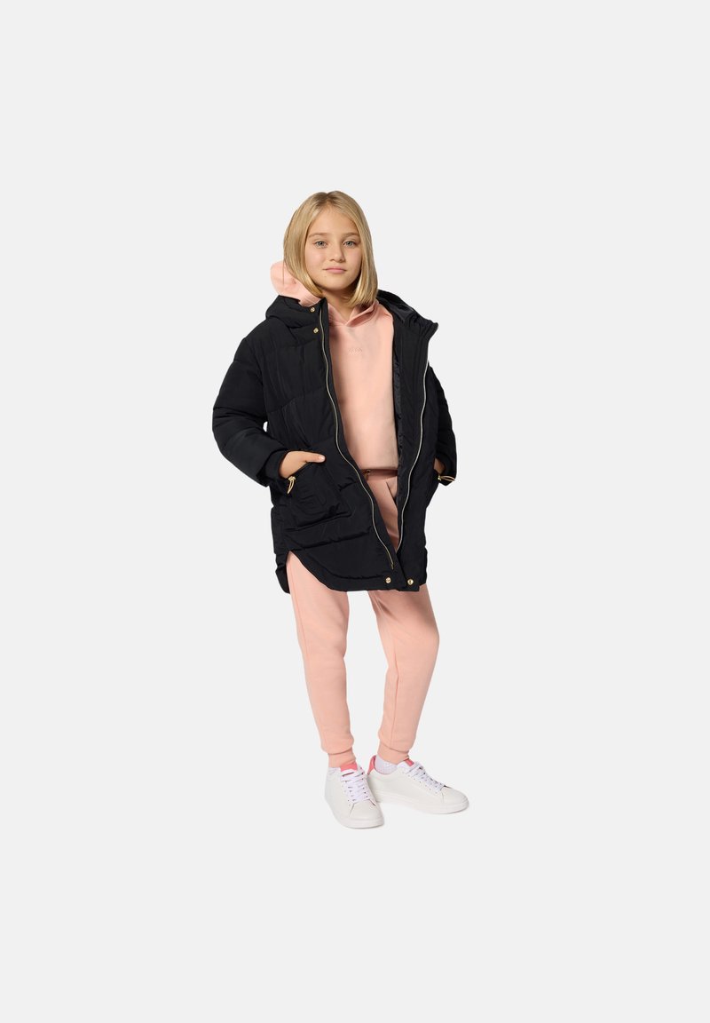 Zwarte pufferjack met capuchon, rits aan de voorkant en zakken, gedragen over een lichtroze hoodie en bijpassende roze trainingsbroek, gecombineerd met witte sneakers.