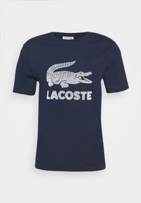 Námořnické modré tričko s krátkým rukávem s velkým bílým grafickým motivem krokodýla a nápisem "LACOSTE" vytištěným pod ním na přední straně.