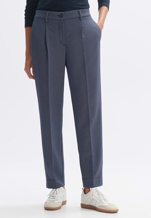 Personne portant un pantalon bleu marine tailleur avec ourlets retroussés, une main dans la poche, associé à des baskets blanches, debout devant un fond uni.