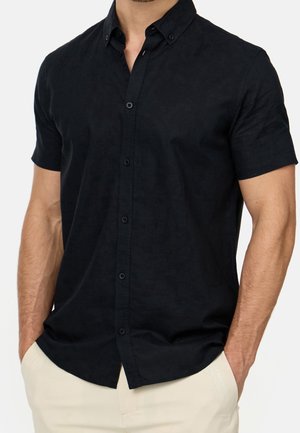 Mężczyzna w czarnej koszuli z krótkim rękawem i guzikiem, włożonej do jasnobeżowych spodni, z rękami w kieszeniach, kadrowany na wysokości szyi i ud.