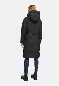 Sort pufferjakke med hætte, der har et quiltet design og vandret syninger. Jakken går ned til midt på læggen og er parret med blå jeans og støvler.