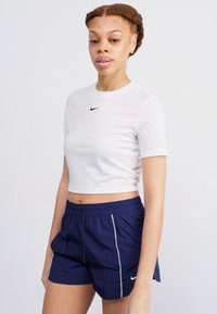 Balta trumpa marškinėliai su trumpomis rankovėmis ir juodu Nike logotipu, derinami su tamsiai mėlynomis šortais su baltais šoninės detalėmis ir elastinga juosta.