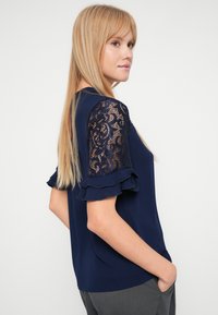 Námornícky modrá blúzka s čipkovými krátkymi rukávmi a volánikmi; hladká látka; voľný strih s okrúhlym výstrihom a jemnými detailmi.