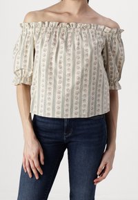 Blouse épaules dénudées en tissu crème avec des motifs floraux verticaux, col froncé et manches bouffantes, assortie à un jean bleu foncé.