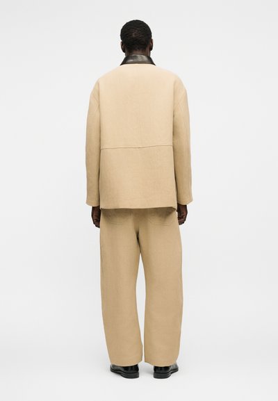 Άνδρας με την πλάτη γυρισμένη, φοράει μπεζ oversized σακάκι και ταιριαχτό παντελόνι με ευρύ πόδι, με μαύρα παπούτσια σε απλό λευκό φόντο.