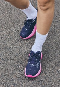 Baskets de sport navy avec des accents roses, tige en mesh, bout arrondi et lacets. Portées avec des chaussettes blanches sur une surface texturée.