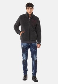 Grijze fleece jas met een hoge kraag, ritssluiting en een gestructureerde afwerking; gecombineerd met versleten blauwe jeans met verfspetters.