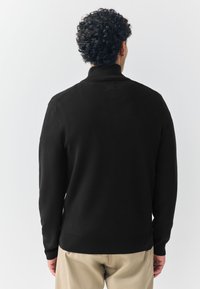 Zwarte gebreide trui met een hoge hals, lange mouwen en geribbelde afwerking bij de manchetten en zoom. Gladde textuur en minimalistisch ontwerp.