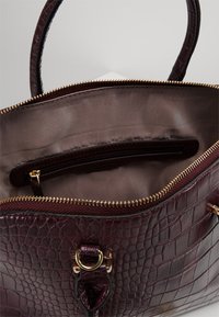 Sac à main en cuir imitation crocodile bordeaux foncé avec fermeture éclair dorée, détail en anneau et intérieur en tissu beige avec poche zippée.