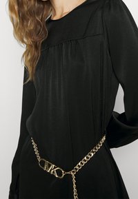 Czarna sukienka z długimi rękawami, z detalami marszczenia przy dekolcie. Udekorowana złotym pasek łańcuszkowym i sprzętem z logo.