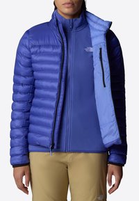 Giacca isolante blu con design a righe, materiale liscio, zip completa e tasche laterali, indossata sopra un interno in pile.