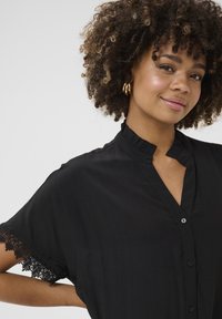 Melna krekls ar viļņotu apkakli, īsām piedurknēm un sarežģītu mežģīņu apdari pie piedurknēm. Iekļauj pogājamu priekšpusi un mīkstu tekstūru.