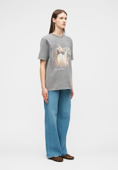 Γυναίκα που στέκεται φορώντας ένα ξεθωριασμένο γκρι μπλουζάκι με στάμπα αλόγου και κείμενο, φαρδιά μπλε τζιν και φλατ παπούτσια με λεοπάρ μοτίβο σε λευκό φόντο.