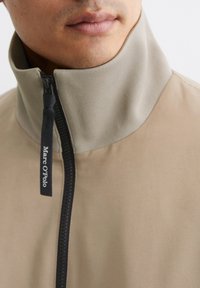 Beige Jacke mit hohem Kragen, ausgestattet mit einem schwarzen Reißverschluss und einem Etikett, das "Marc O'Polo" liest. Glatte Stofftextur mit einem minimalen Design.