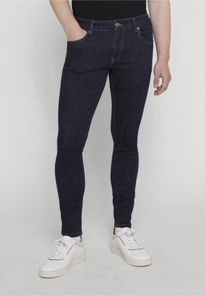 Asmuo dėvi tamsiai mėlynus siaurus džinsus ir baltus sportinius batelius, stovi su viena ranka kišenėje priešais vienspalvį foną.