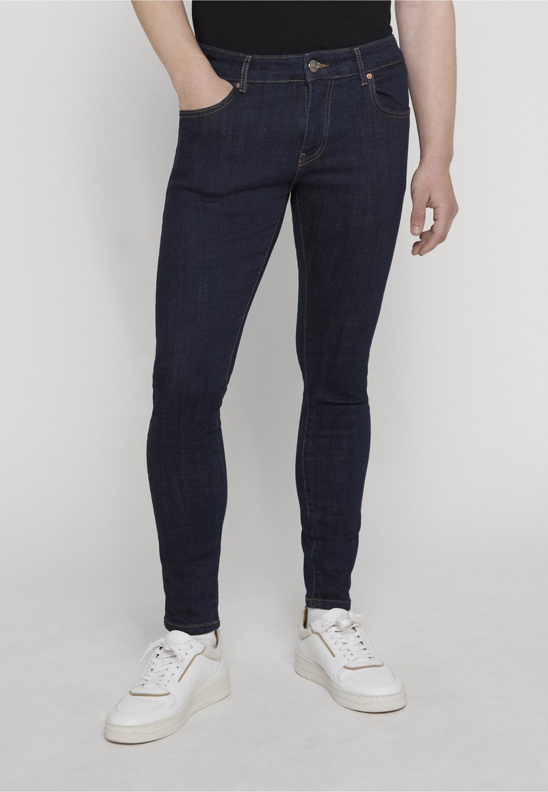 Personne portant un jean skinny bleu foncé et des baskets blanches, debout avec une main dans la poche sur un fond uni.
