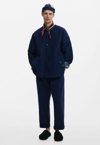 Giacca trapuntata blu navy con chiusure a pressione, tasche laterali e una camicia a fantasia floreale sotto. Abbinata a pantaloni blu navy e pantofole nere.
