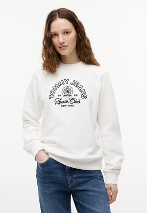 Młoda kobieta o falowanych włosach nosi białą bluzę Tommy Jeans z napisem "Sports Club New York" i niebieskie dżinsy, stoi z ręką w kieszeni.