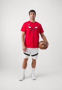 Rød Nike T-shirt med Chicago Bulls-logo, hvide shorts med en sort stribe, holder en basketball. Hvide sneakers fuldender looket.
