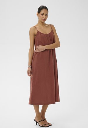 Femme debout portant une robe midi sans manches couleur rouille avec de fines bretelles et des sandales à talons noires, sur un fond clair uni.