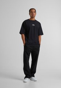 Sort bomulds oversized t-shirt med et hvidt grafisk logo på brystet, parret med sorte bukser og hvide sneakers.