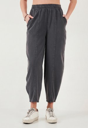 Személy laza sötétszürke, gumis derék- és szárbefejezésű nadrágban, kezét zsebében tartva, fehér sportcipővel párosítva.