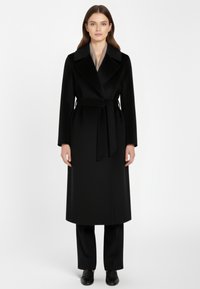 Vrouw in een lange zwarte, met een riem gecentraliseerde jas over zwarte broek en zwarte schoenen, staand tegen een effen witte achtergrond.