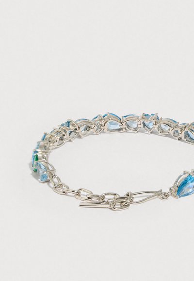Pulseira de prata com pedras preciosas azuis em formato de lágrima, ligadas por correntes de metal, com um fecho decorativo numa das extremidades.