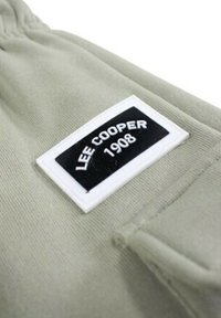Étiquette Lee Cooper en noir et blanc, cousue sur un fond en tissu vert clair ; forme rectangulaire aux bords arrondis et surface texturée.