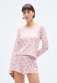 Σετ πυτζάμες σε ανοιχτό ροζ χρώμα, με μπλούζα μακρυμάνικη και στρογγυλή λαιμόκοψη, διακοσμημένη με σχέδιο από φιόγκους, συνοδευόμενη από σορτς με ριγέ κορδόνι στη μέση.