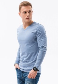 Světle modrá dlouhá halenka s výstřihem do V, která se vyznačuje jemnou texturou a přiléhavým střihem, v kombinaci s roztrhanými modrými džíny.