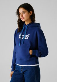 Haut à capuche bleu marine avec une poche frontale, arborant le texte blanc "Red Bull RACING" et un cordon de serrage turquoise. Tissu doux avec une coupe ample.