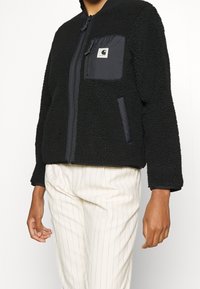Czarna polarowa kurtka na zamek z kontrastową kieszenią z czarnego materiału i akcentami z tkaniny, z teksturowaną powierzchnią i luźnym fasonem.