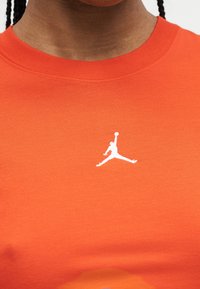 Κοντινό πλάνο πορτοκαλί πουκάμισου με μικρό λευκό λογότυπο Jumpman στο κέντρο του στήθους, που φοριέται από άτομο με πλεξούδες.