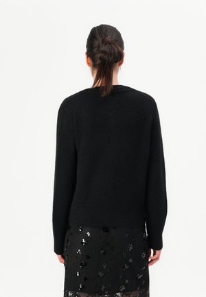 Femme aux cheveux foncés attachés en chignon porte un pull noir à manches longues et une jupe noire avec des motifs floraux en sequins, vue de dos.