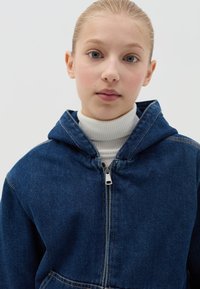 Sudadera de mezclilla con cremallera en la parte frontal, color azul, y bolsillo tipo canguro. Lleva puesta una camiseta de cuello alto blanca de canalé. Textura suave.
