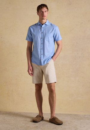 Junger Mann, der ein hellblaues kurzärmeliges Hemd mit Knöpfen, beige Shorts und braune Slipper trägt, steht vor einem beigen Hintergrund.