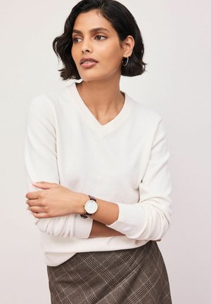 Frau mit kurzen dunklen Haaren trägt einen weißen V-Ausschnittpullover, einen karierten Rock, Creolen und eine Armbanduhr, schaut zur Seite mit verschränkten Armen.