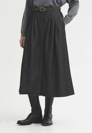 Femme portant une jupe midi gris foncé boutonnée avec des poches, une ceinture noire cloutée, un pull gris et des bottes noires à lacets montant jusqu’au milieu du mollet.