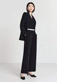 Schwarzer maßgeschneiderter Anzug mit einem einreihigen Blazer, weit geschnittenen Hosen und einem weißen Taillenbund; kombiniert mit schwarzen Lack-Slingbacks.