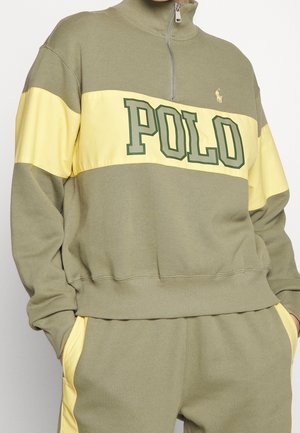 Groen en geel sweatshirt met halve rits en de tekst "POLO" op de borst, gecombineerd met bijpassende broek met gele strepen aan de zijkant en handen in de zakken.