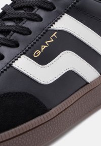 Μαύρο αθλητικό παπούτσι με λευκό κυματιστό σχέδιο, υφασμάτινες λεπτομέρειες σουέντ, μαλακή καφέ ελαστική σόλα και χρυσή λεπτομέρεια λογότυπου στο πλάι.