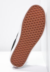 Semelle d'une basket noire et blanche inclinée contre un mur, montrant une semelle à motif gaufré marron sur fond blanc.