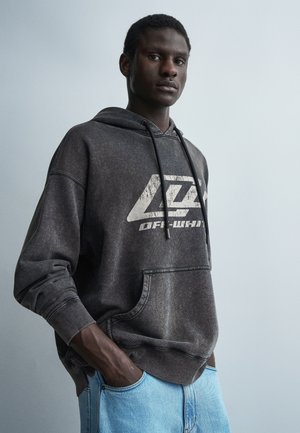 Młody mężczyzna stojący z rękami w kieszeniach, ubrany w wyblakłą czarną bluzę z kapturem z nadrukiem tekstowym oraz jasnoniebieskie dżinsy na jednolitym tle.