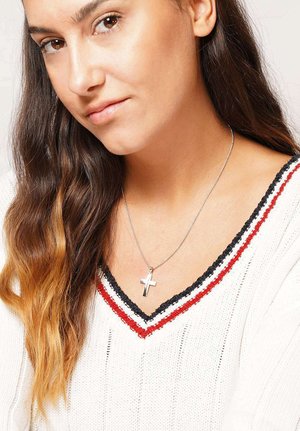 Young woman with long wavy hair wearing a white knit sweater with black and red trim, and a silver cross necklace.