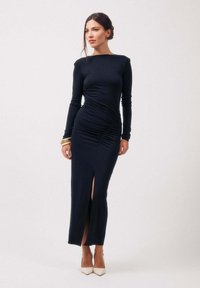 Femme en robe noire longue et ajustée avec une fente sur la cuisse, portant des talons blancs et des bracelets dorés, debout devant un fond clair uni.