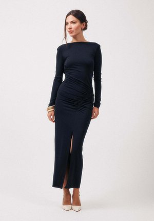 Femme en robe noire longue et ajustée avec une fente sur la cuisse, portant des talons blancs et des bracelets dorés, debout devant un fond clair uni.